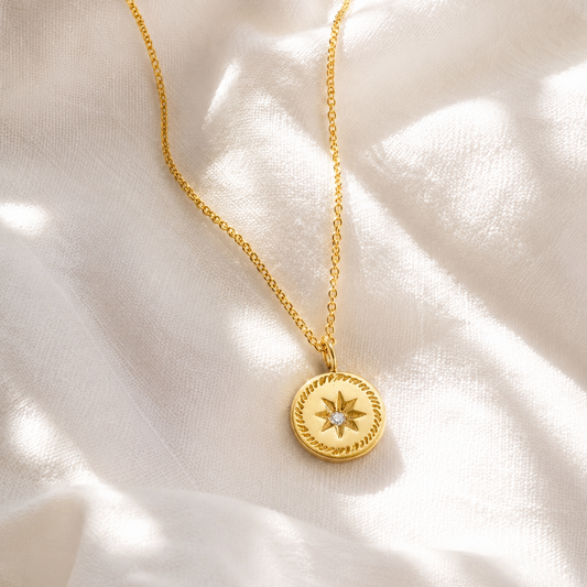 A gold chain necklace with a round pendant featuring a star motif and a small clear gemstone, displayed on draped white fabric.