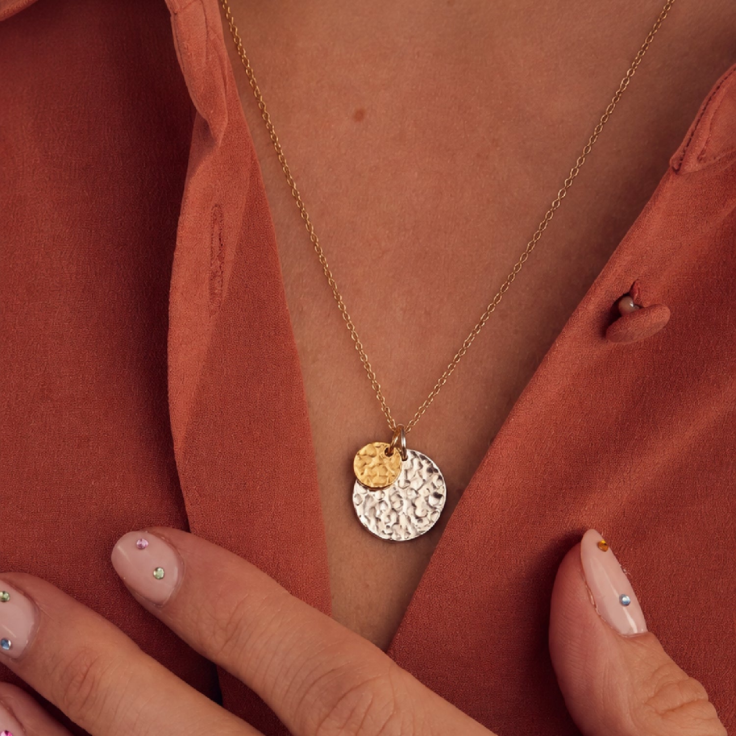 A close-up of a person wearing a delicate gold chain necklace with two round disc pendants, one textured silver and one smooth gold, over a rust-orange satin shirt.